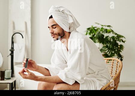 Young metrosexual man in bathrobe sitting on chair in bathroom and taking care of his hands Stock Photo