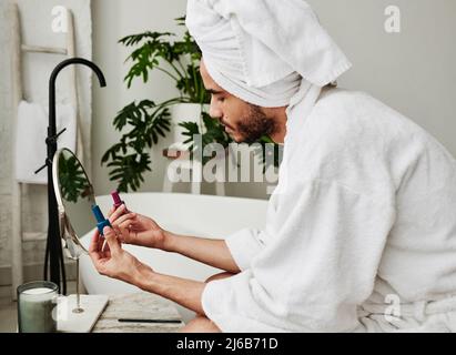 Young handsome man in bathrobe choosing trendy colour of varnish for his manicure while sitting in bathroom Stock Photo