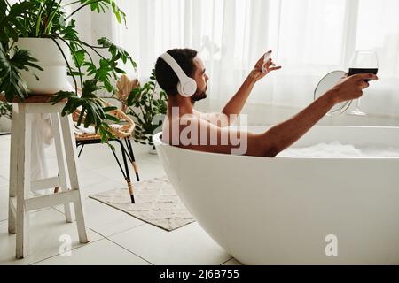 Young man relaxing in bathtub with glass of red wine and enjoying music in headphones during spa procedure in bathroom Stock Photo