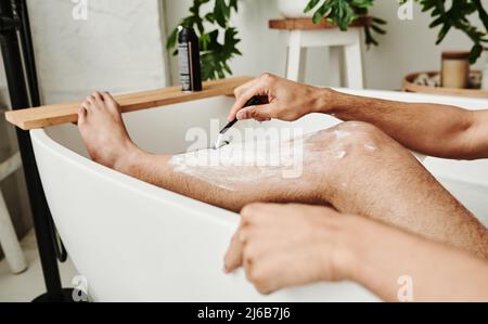 Close-up of young man using razor to shave his legs while sitting in bath Stock Photo