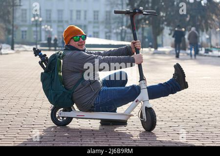 Guy photographer sits on an electric scooter in the spring in the park. He is wearing a warm jacket and hat. Nearby lies snow and cold Stock Photo