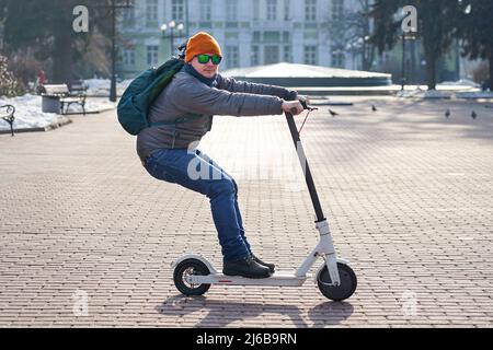 The guy rides an electric scooter in the spring in the park. He is wearing a warm jacket and hat. Nearby lies snow and cold Stock Photo