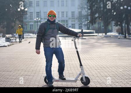 The guy rides an electric scooter in the spring in the park. He is wearing a warm jacket and hat. Nearby lies snow and cold Stock Photo