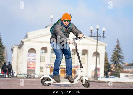 The guy rides an electric scooter in the spring in the park. He is wearing a warm jacket and hat. Nearby lies snow and cold Stock Photo