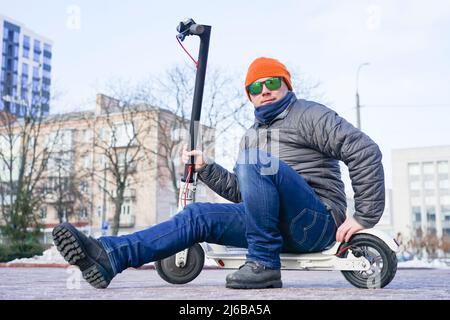 The guy sits on an electric scooter in the spring in the park. He is wearing a warm jacket and hat. Nearby lies snow and cold Stock Photo