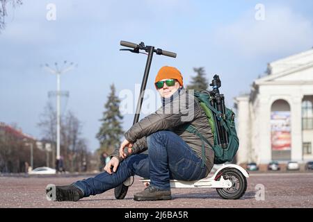 Guy photographer sits on an electric scooter in the spring in the park. He is wearing a warm jacket and hat. Nearby lies snow and cold Stock Photo
