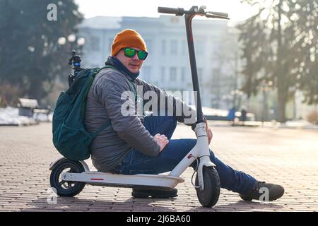 Guy photographer sits on an electric scooter in the spring in the park. He is wearing a warm jacket and hat. Nearby lies snow and cold Stock Photo