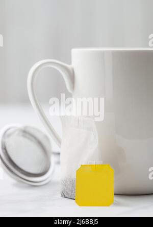 Teabag with yellow blank tag of black tea next to porcelain cup on white background with infuser strainer. Stock Photo