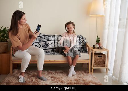 Mother photographing daughter with miniature poodle sitting on couch at home Stock Photo