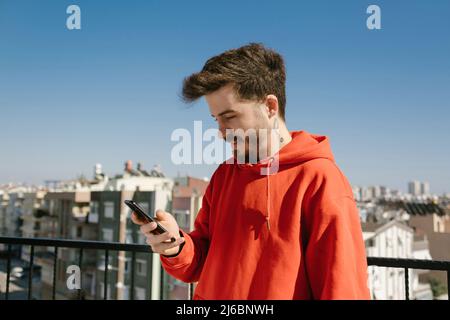 Handsome young man wearing red hoodie using phone. Man looking to smartphone screen with smiles. Messaging with friend or watching video or scrolling Stock Photo