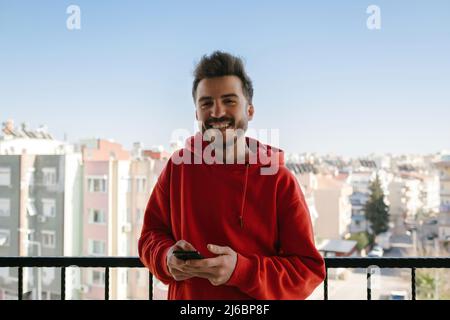 Young happy man wearing red hoodie excited cheerful joyful delightful curious guy sending and getting messages to his lover. Using phone. Looking to c Stock Photo