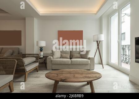 Living room with oval unvarnished wooden table on wooden floors, balcony with metal railings and gray fabric and rattan sofas Stock Photo