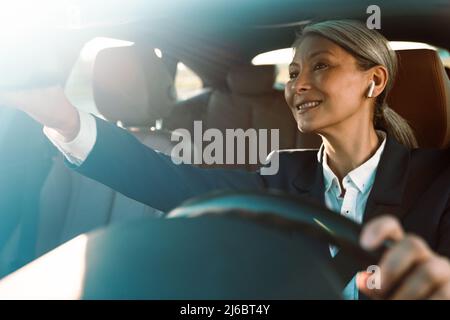 Mature asian woman using earphones and looking in rear-view mirror while driving car Stock Photo