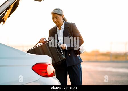 Mature asian businesswoman wearing suit putting her suitcase in trunk Stock Photo