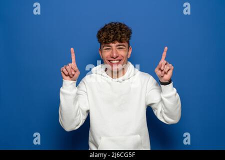 Portrait of a happy young man with curly hair pointing finger up at copy space isolated over blue background. Stock Photo