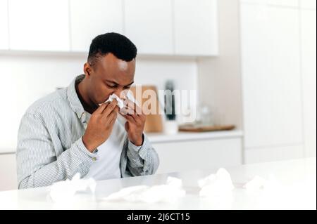 Ill and allergy concept. Unhealthy sick african american man with runny nose. Black guy is sick with flu, he sneezes and blows his nose in a napkin while sitting at home in quarantine alone Stock Photo