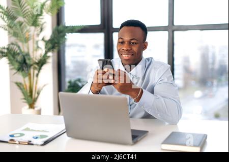 Positive attractive young adult african american business man, agent, manager, sits workplace in modern office, uses smartphone, chatting on the phone with colleagues or clients, smiling friendly Stock Photo