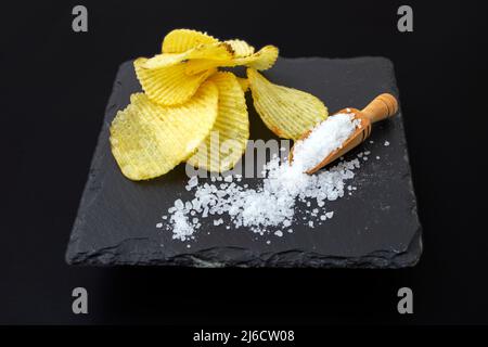 Potato chips stack nest to a scoop full of sea salt, on a black stone tile, close up Stock Photo