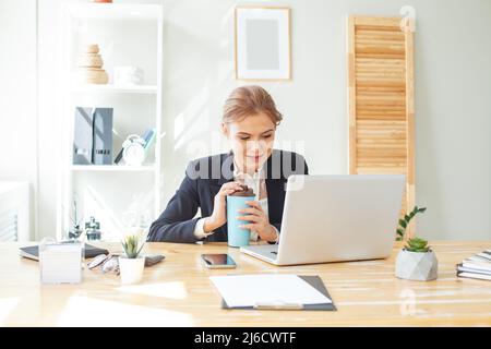 Happy blonde businesswoman using laptop and drinking coffee in office Stock Photo