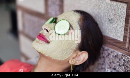 A beautiful woman with facial clay mask on face and fresh cucumber on eyes relaxing at Home. Portrait of beautiful spa girl. Beauty, healthcare. Multa Stock Photo