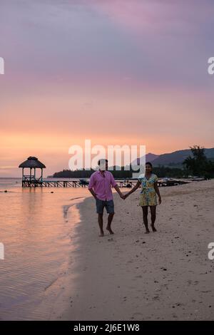 couple watching the sunset on a tropical beach with a wooden pier in the ocean, men and woman sunset on the beach during luxury vacation.  Stock Photo
