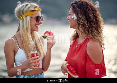 Girlfriends talking and enjoying a glass of beer and piece of watermelon fruit by sea. Togetherness, holiday, friendship, lifestyle concept. Stock Photo