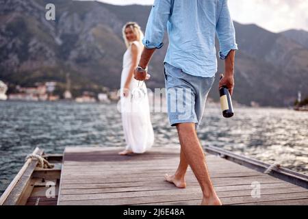 Groom with bootle of wine and glasses, romantic date by seaside. Newlywed coupe on honeymoon. Wedding, honeymoon, love concept Stock Photo