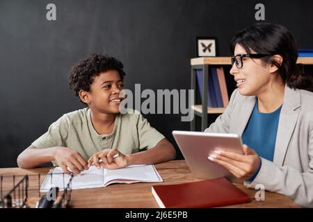Smiling Latin teacher in glasses sitting at table and using tablet while assisting boy with homework at tutorial Stock Photo