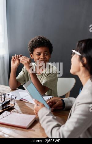 Smiling cute Black student boy sitting at table with workbook and twisting pen while talking to tutor Stock Photo
