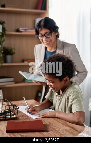 Latin tutor with tablet standing at African American schoolboy who doing task during individual lesson Stock Photo