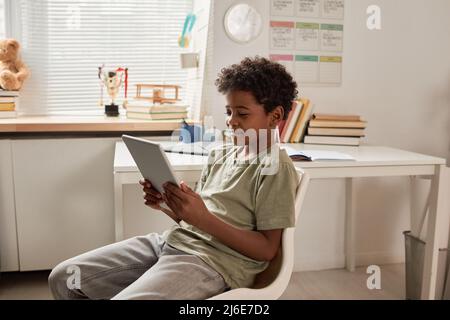 Smiling African American boy with curly hair sitting on chair and using tablet in own room Stock Photo