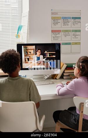 Rear view of multi-ethnic children sitting at table with modern computer and listening to Hispanic tutor online Stock Photo