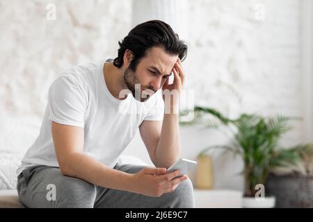 Upset young man using smartphone in bed Stock Photo