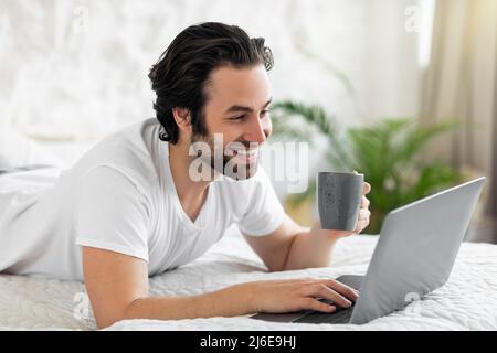 Young millennial guy using laptop in bed, drinking coffee Stock Photo