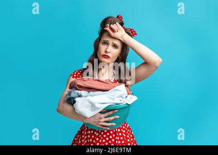 Tired young pinup woman in retro style dress holding clothes for washing or ironing, wiping forehead on blue background Stock Photo