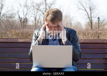 A tired blond man of 40-45 years old clutched his head in pain while working computer while sitting on a bench. Stock Photo