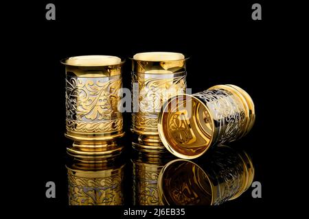 Three Golden Antique Vintage Brass Shot Glass Gilded on black background. metal Wine Cup goblet with Carving Engraving pattern. Stock Photo