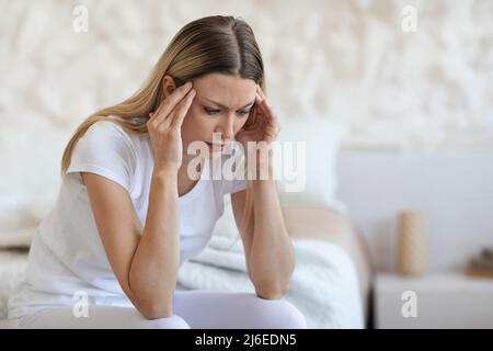 Unhappy young lady sitting on bed, rubbing her temples Stock Photo