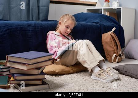 Concentrated pretty girl sitting on pillow and making notes in workbook while doing homework Stock Photo