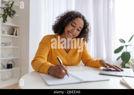 Young black woman studying or working online with laptop at home, taking notes, enjoying distance job or education Stock Photo