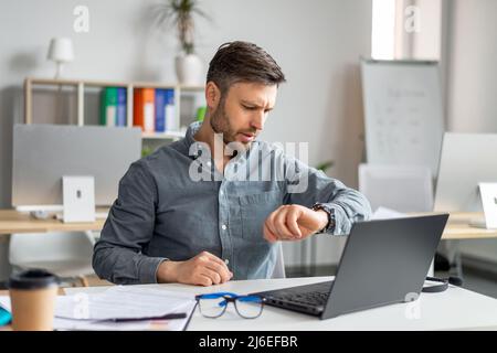 Business career and time management. Mature businessman using laptop and checking time, looking at watch Stock Photo