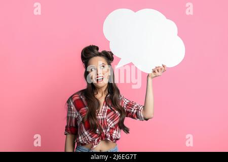 Young pinup woman in retro outfit holding blank white speech bubble on pink studio background Stock Photo