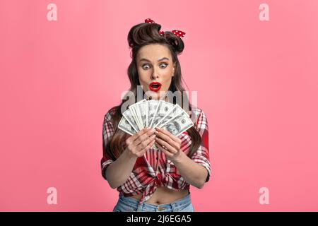 Excited millennial pinup lady in retro style wear holding fan of money, winning online casino bet or lottery Stock Photo