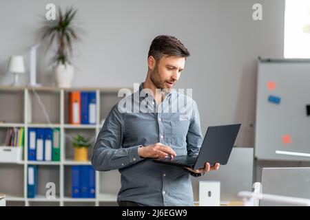 Handsome mature businessman using laptop computer, working online at modern office, typing on keyboard Stock Photo