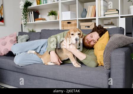 Young bearded man lying on sofa and embracing his dog during leisure time at home Stock Photo