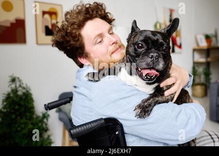 Young bearded man sitting in wheelchair and holding his french bulldog on his arms, he playing with his pet Stock Photo