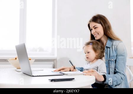 Stay home. Mother works from home, she sits in the kitchen and using laptop for remote work, her cute little girl sits nearby on a chair and looks at Stock Photo