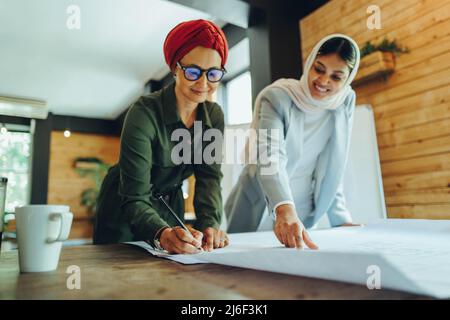 Happy Muslim architects working on blueprint drawings in a modern office. Two creative businesswomen planning a new innovative project. Female designe Stock Photo
