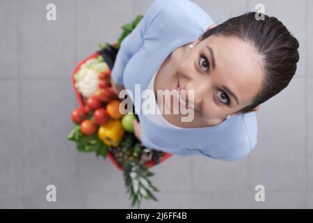 Young woman holding a shopping basket full of fresh vegetables and fruits, she is smiling at camera, top view Stock Photo