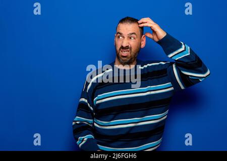 Young doubtful man thinking, scratching his head trying to find solution, isolated on blue background. Stock Photo